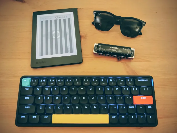 A Photograph of a computer keyboard, sunglasses, a harmonica, and an ebook reader.